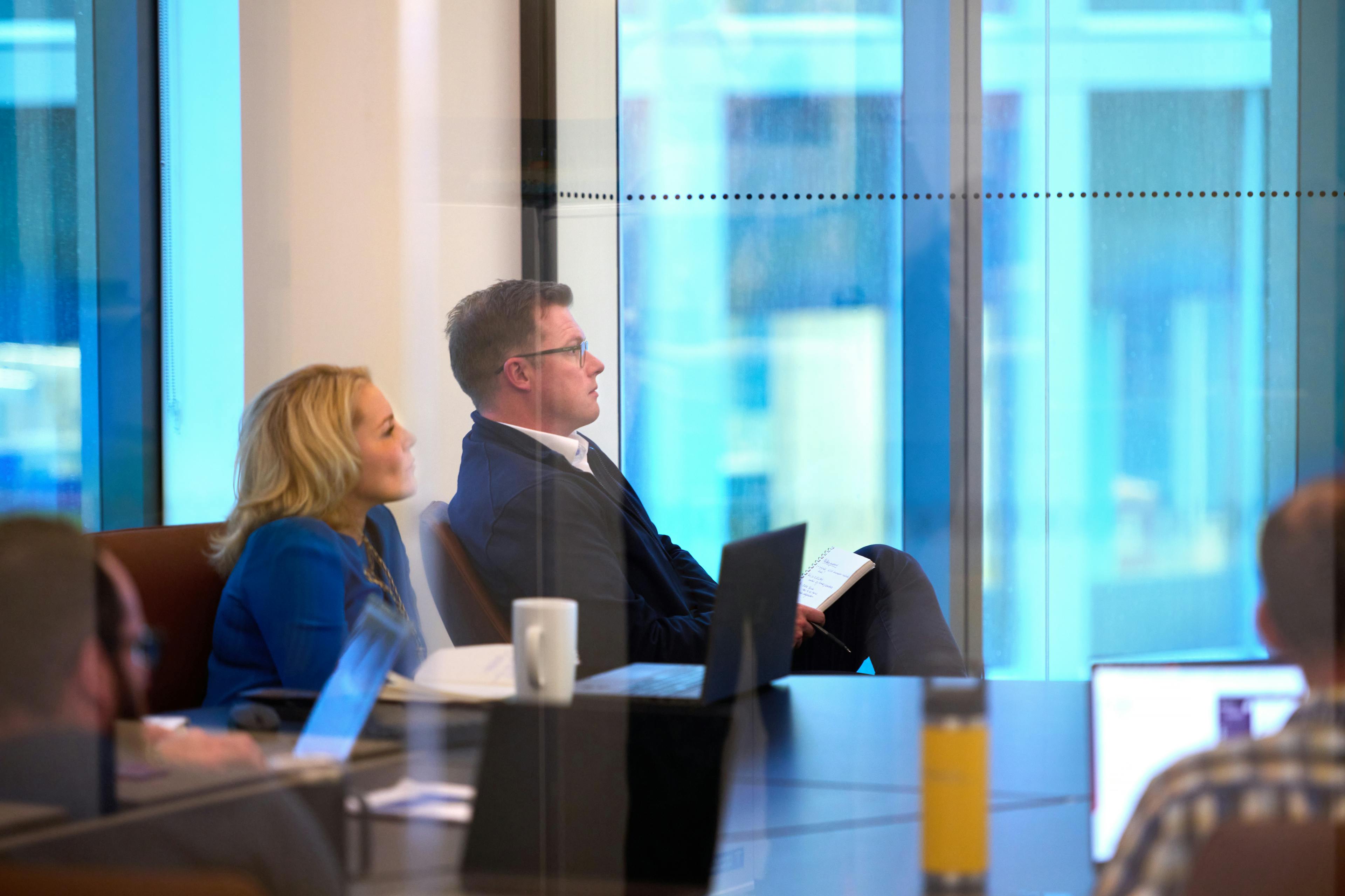 Business leaders are seated at a conference room table, looking intently off-camera