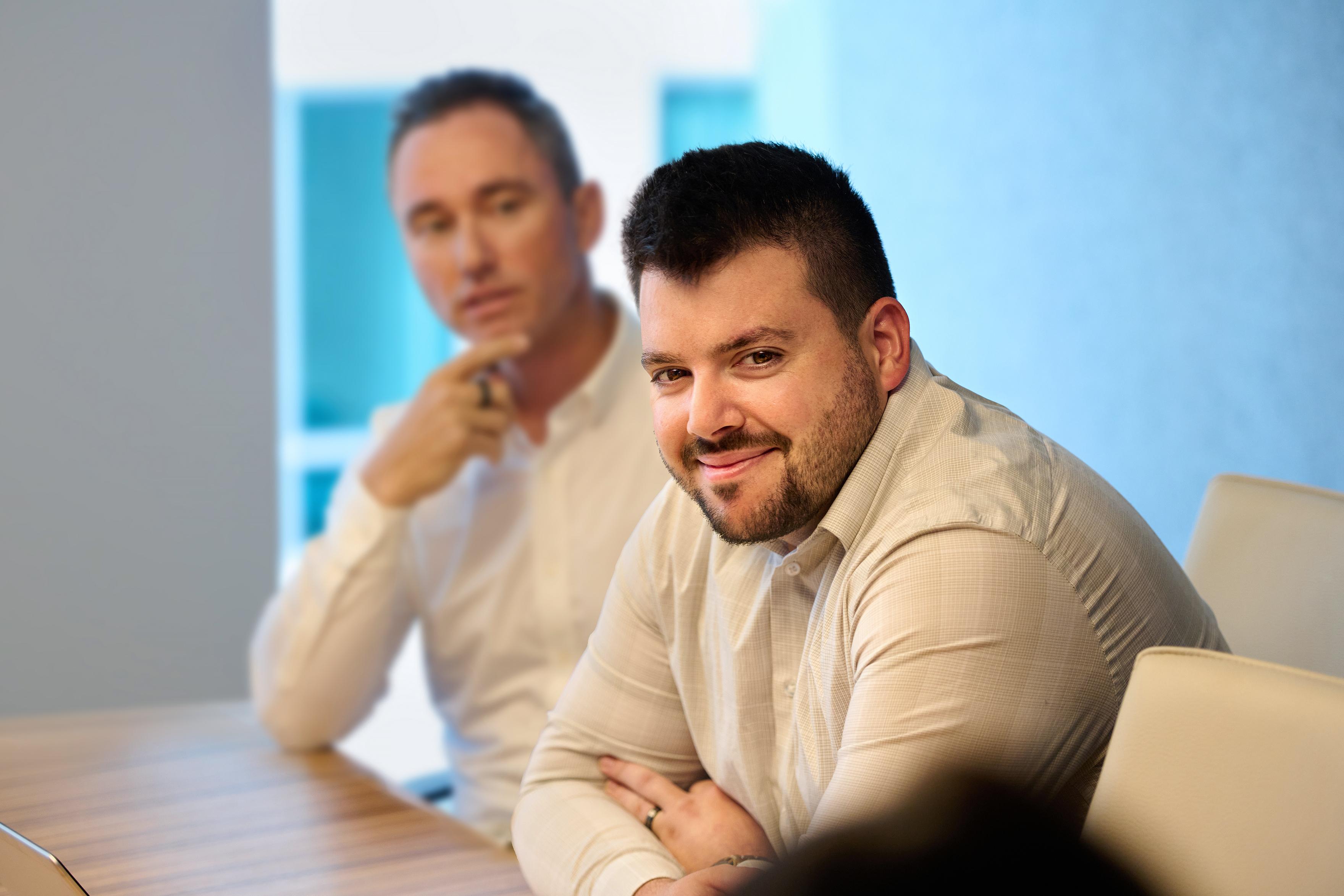 A sales employee smiles confidently at the camera. In the background, another employee looks thoughtfully at something in the distance